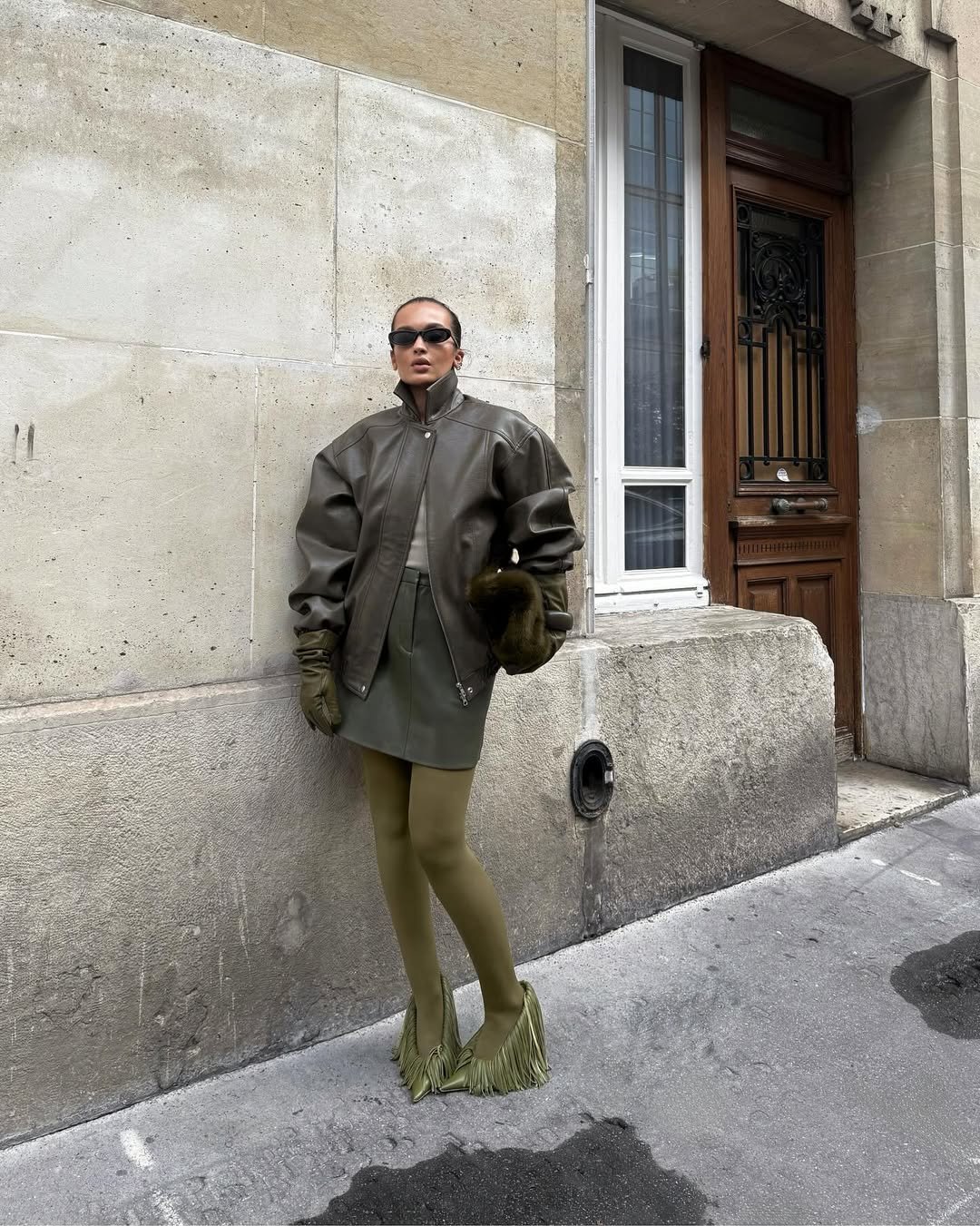 Modelo posa com jaqueta oversized de couro marrom com gola funil alta, saia curta, luvas e meias verdes, em cenário urbano