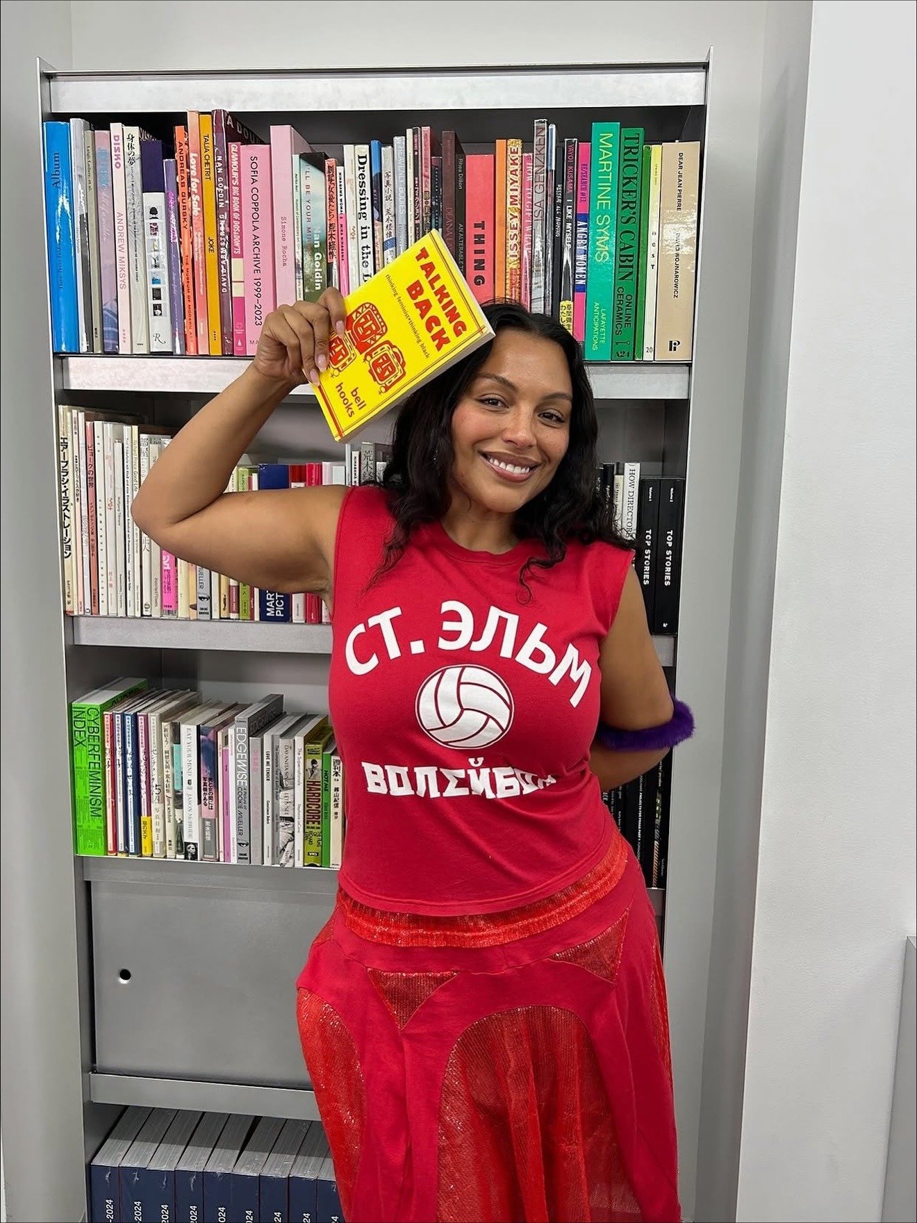 Paloma Elsesser posa sorridente com regata vermelha de estampa russa e saia vermelha translúcida, segurando livro amarelo.