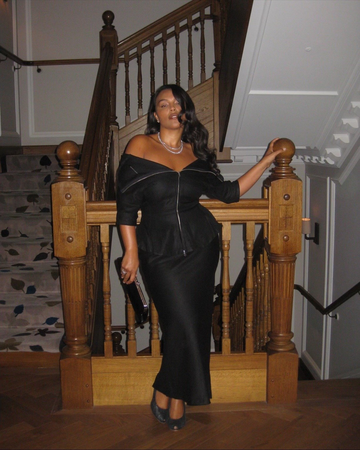 Paloma Elsesser posa com vestido preto justo de tecido acetinado, decote ombro a ombro e zíper frontal, em ambiente elegante.