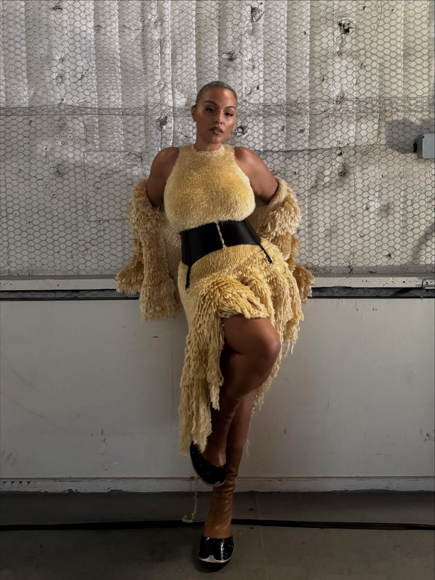 Paloma Elsesser posa confiante com vestido e casaco de textura felpuda amarela, cinto preto marcando a cintura e botas