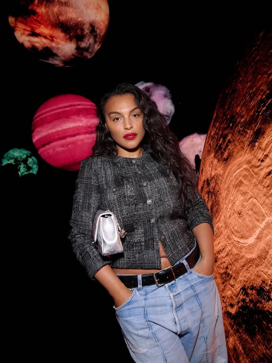 Paloma Elsesser posa confiante com jaqueta cropped texturizada preta, jeans claros, cinto preto e bolsa prateada em cenário