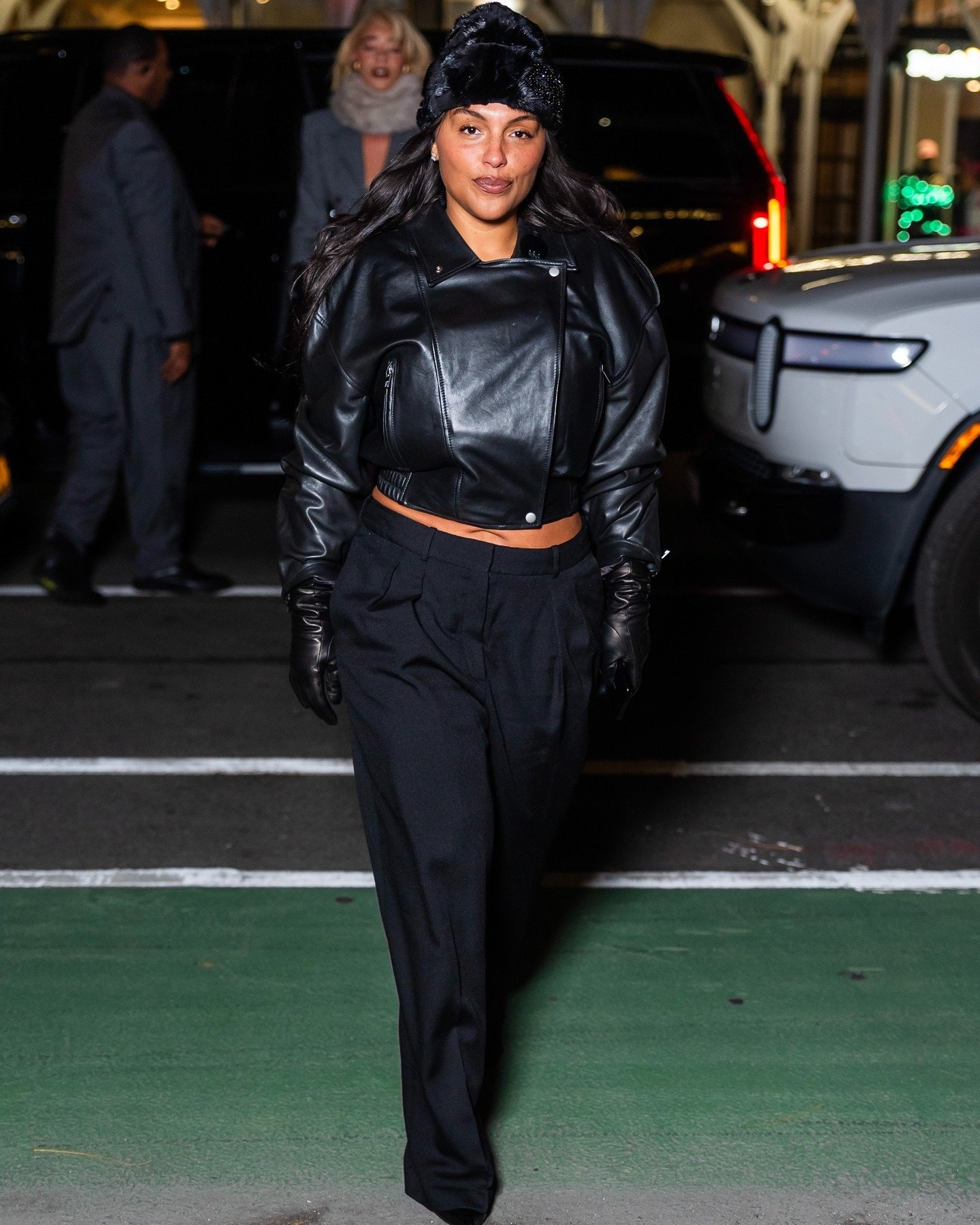 Paloma Elsesser posa confiante com jaqueta cropped de couro preto, calça ampla preta, luvas de couro e touca felpuda preta.