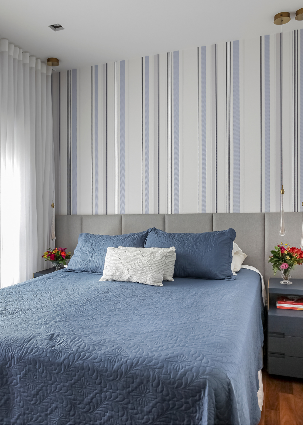 Quarto com papéis de parede listrados em tons de branco e azul, cama com colcha azul texturizada e clima acolhedor.