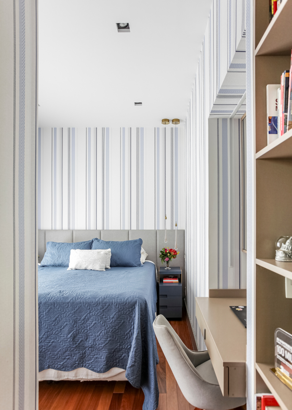 Quarto com papéis de parede listrados em tons de branco e azul, cama com colcha azul texturizada e ambiente clean e