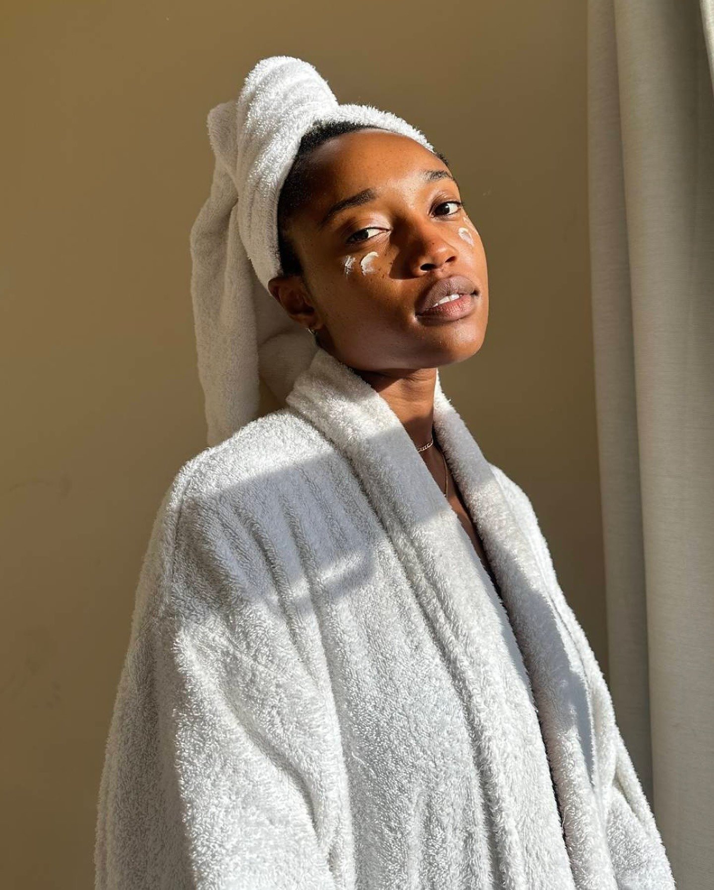 A imagem retrata uma mulher com pele radiante, usando um roupão branco e com uma toalha na cabeça. Ela aplica um creme no rosto, destacando a importância do cuidado com a pele.