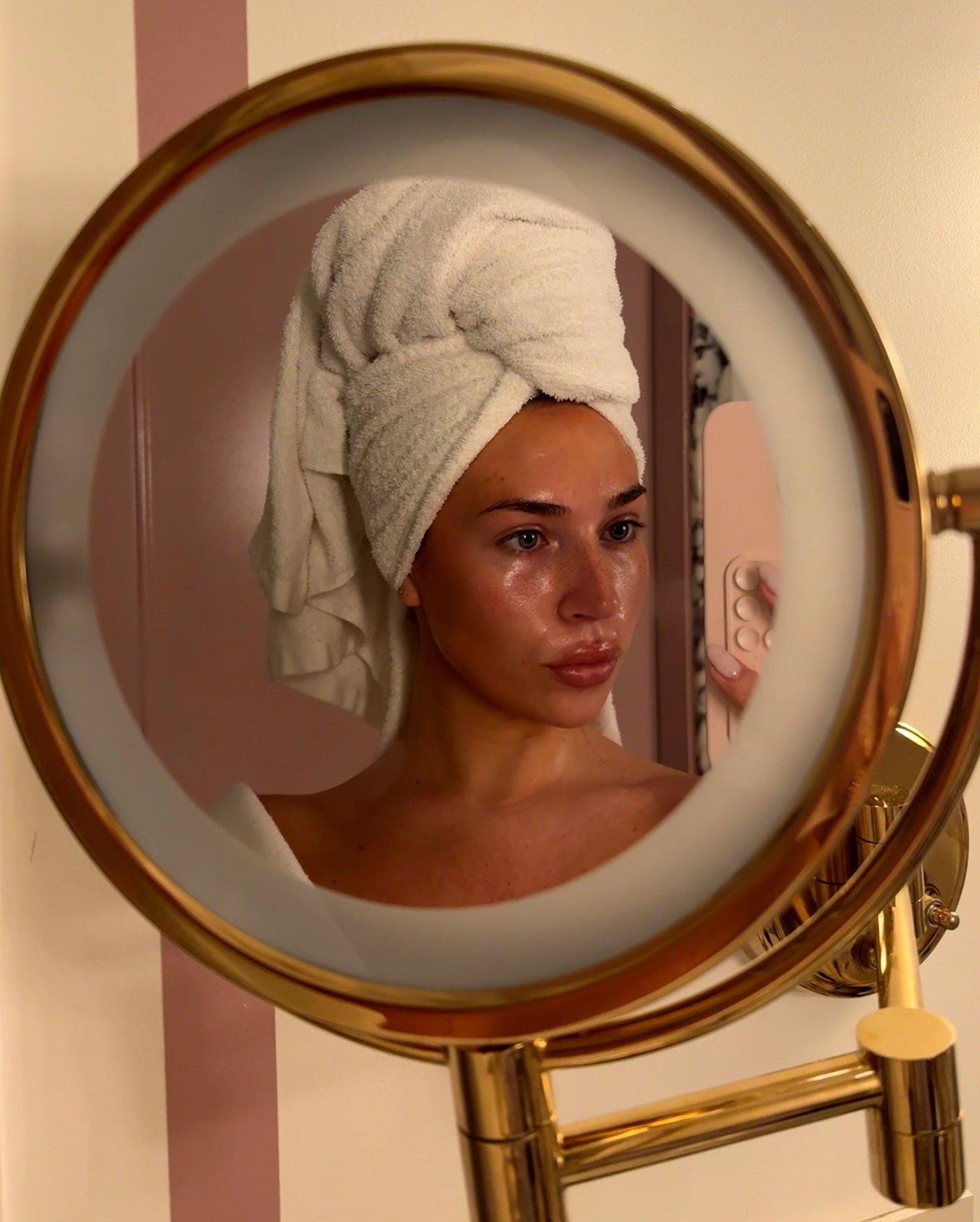 A imagem retrata uma mulher com cabelo preso em uma toalha branca, olhando para um espelho redondo dourado. Sua pele apresenta um brilho saudável, sugerindo o uso de produtos de beleza, como ácido hialurônico.