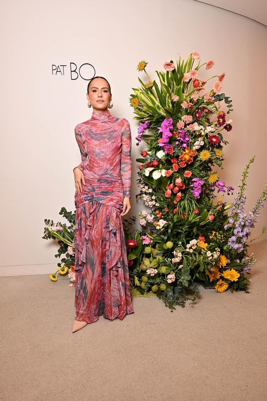 Modelo em vestido longo da PatBO, com estampa vibrante e detalhes em babados, posando ao lado de um arranjo floral exuberante. A paleta de cores do vestido complementa as flores, destacando a estética moderna e romântica da coleção.