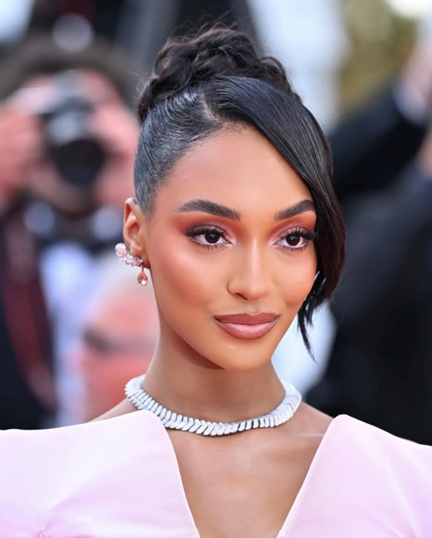 Penteado chique com coque alto trançado e franja lateral lisa, realçado por maquiagem em tons terrosos e colar branco