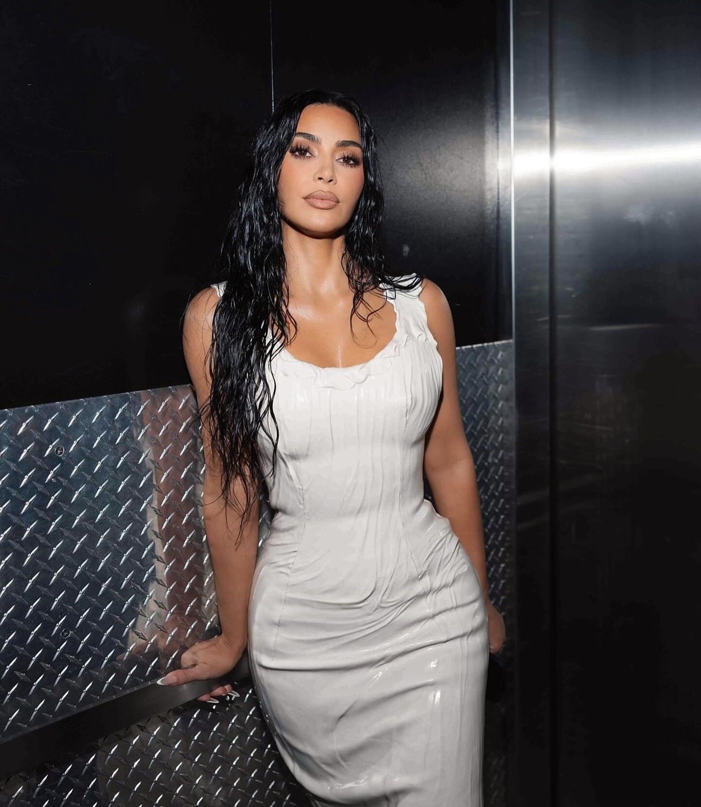 Modelo posa com penteado chique de cabelos longos e molhados, vestido branco justo de tecido brilhante em cenário urbano