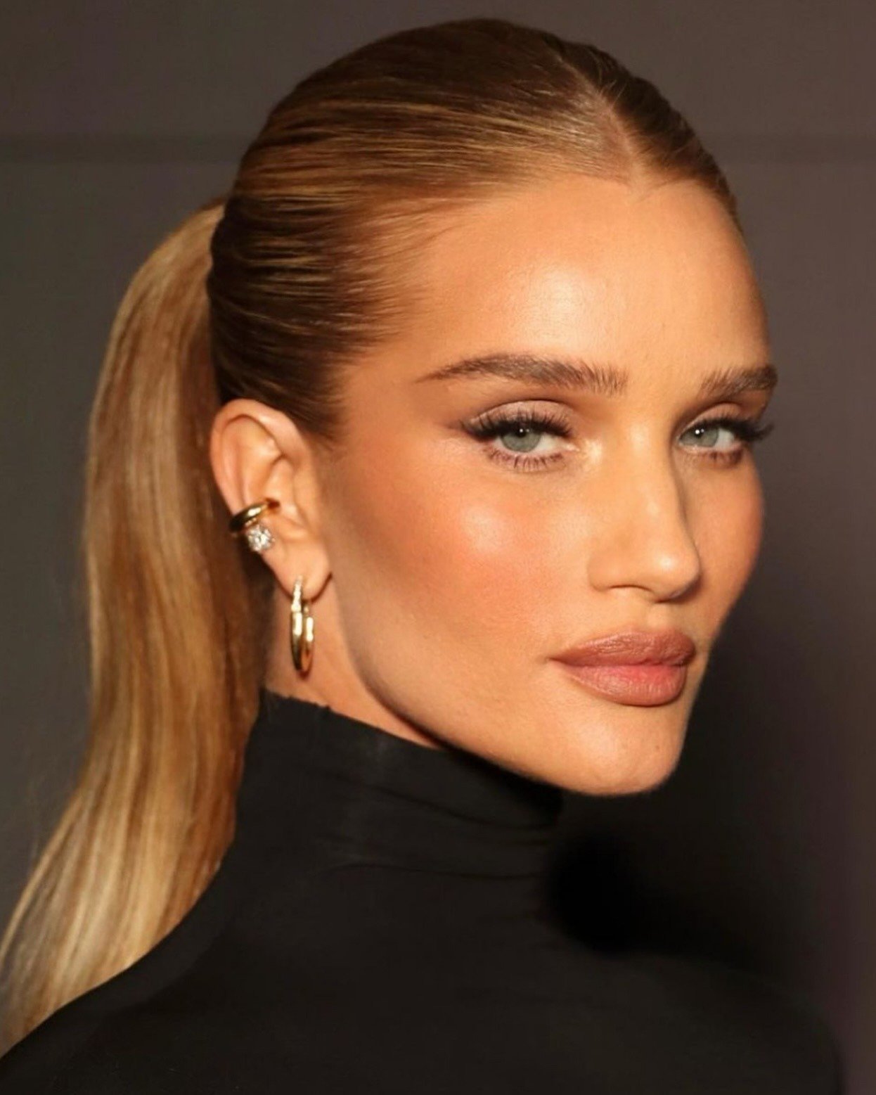 Penteado chique com rabo de cavalo liso e alto, maquiagem neutra e look elegante em gola alta preta.