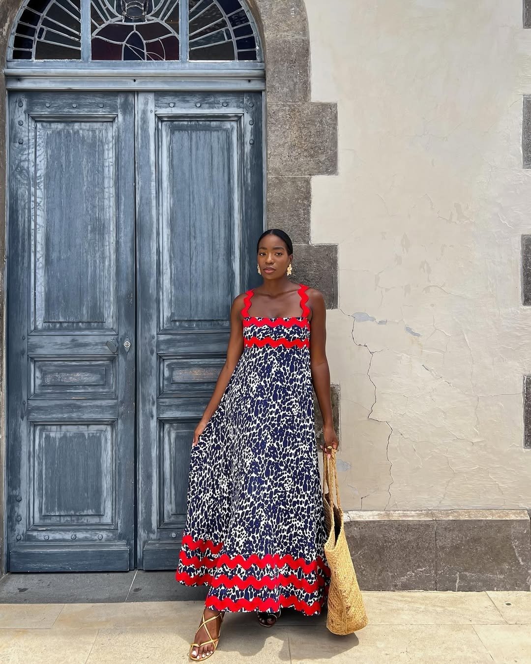 A rasteirinha dourada brilha sob a luz, complementando o vestido longo com estampa de manchas em azul marinho e detalhes vermelhos. O look é perfeito para dias quentes, trazendo elegância e conforto.