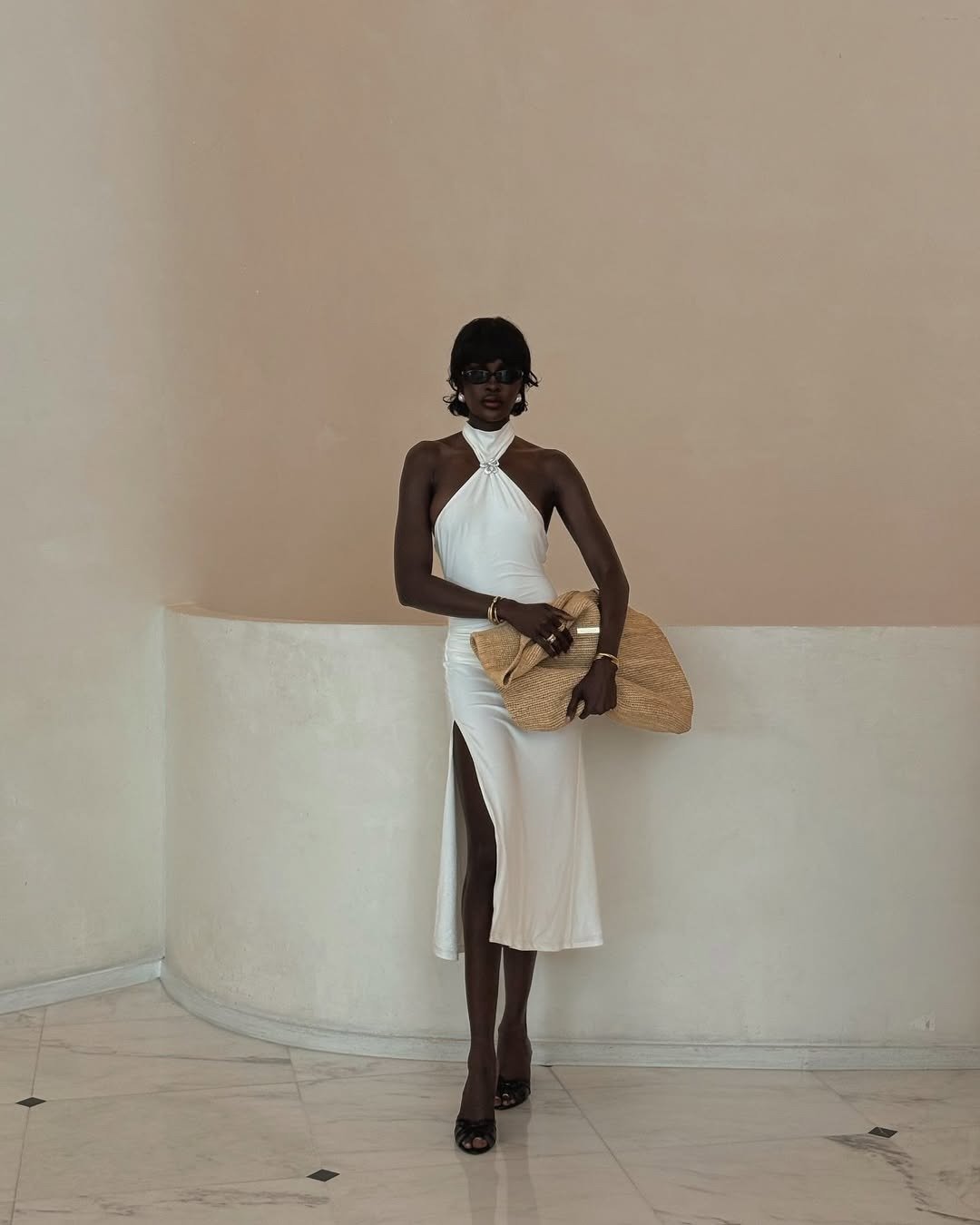 A imagem apresenta uma mulher vestindo um elegante vestido branco com detalhes que destacam a silhueta. O modelo possui um decote halter e uma fenda lateral, conferindo um toque sofisticado.