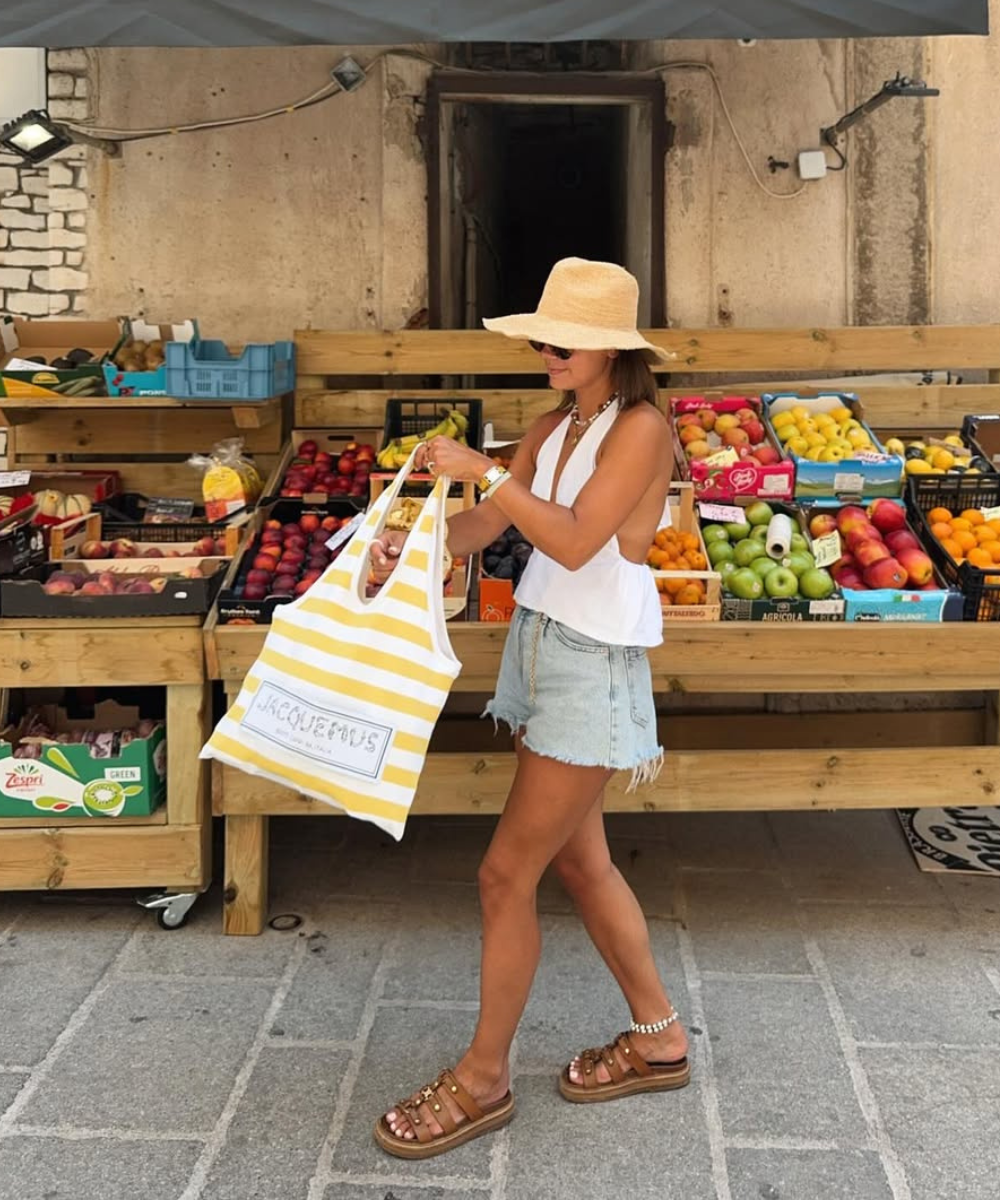Mulher caminha com regata branca de alças finas, shorts jeans desfiados, chapéu de palha e bolsa listrada em clima leve.