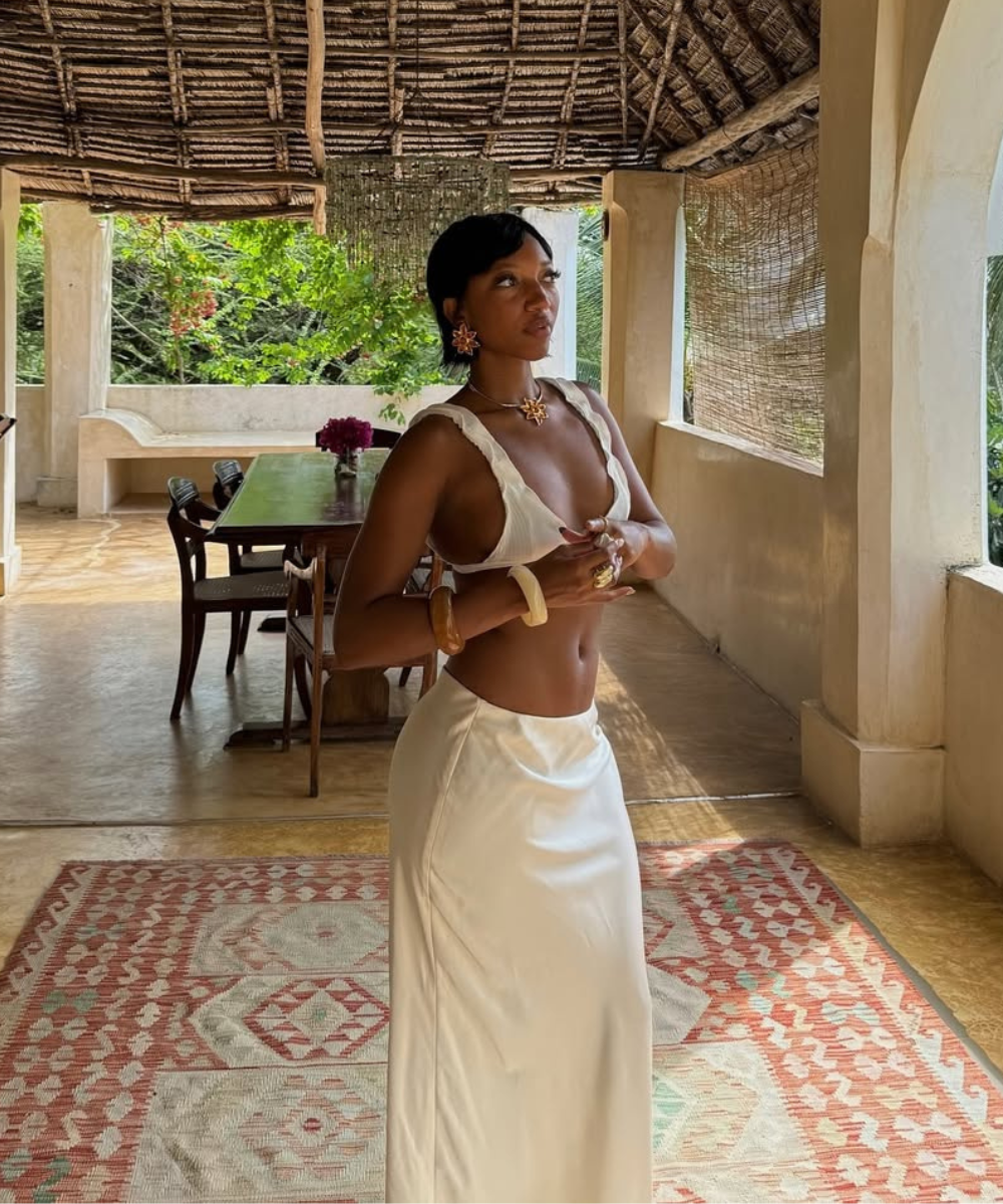 Modelo posa com top branco de tecido leve e saia longa fluida em ambiente rústico, evocando elegância e leveza.