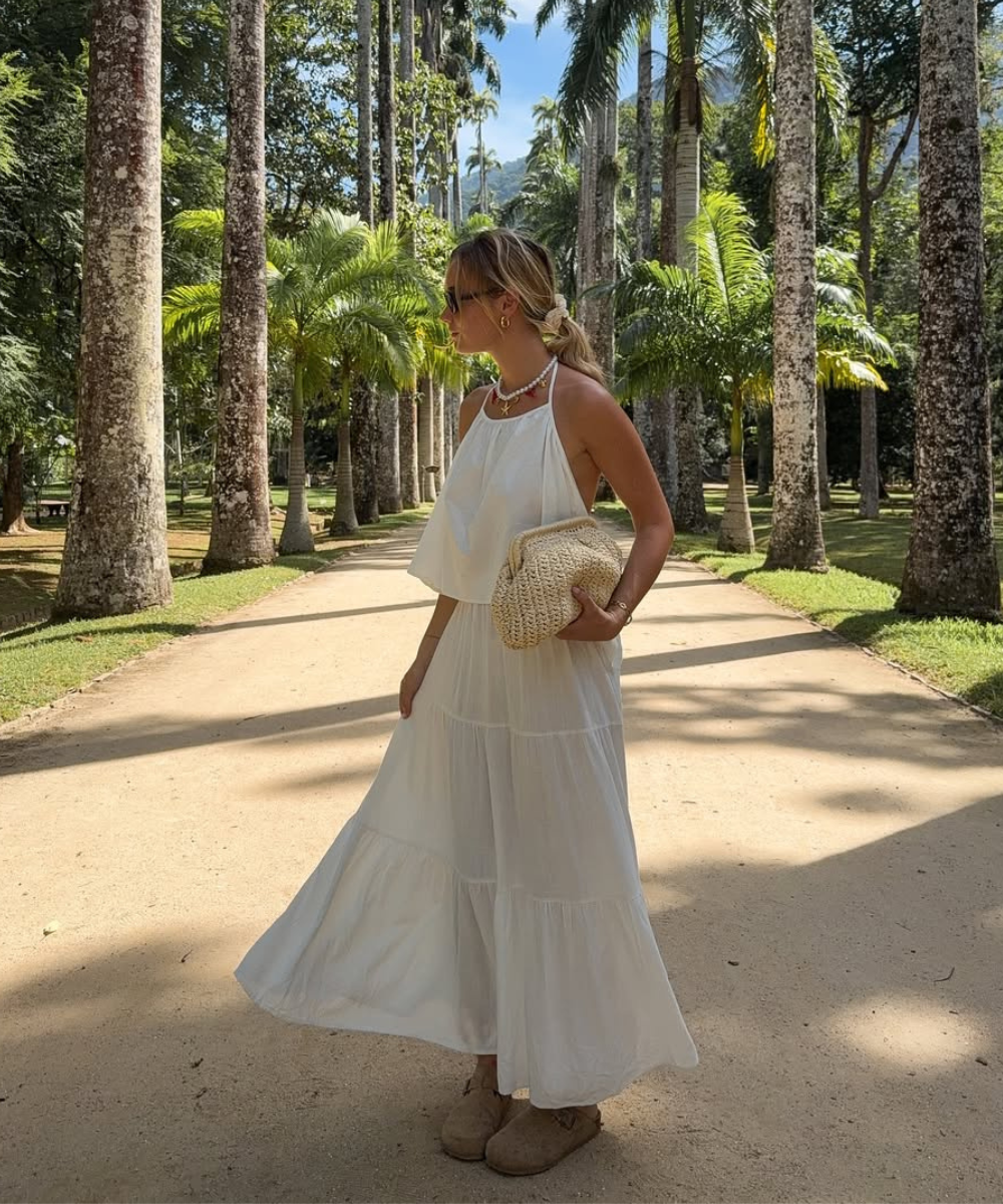 Mulher posa com vestido branco fluido de tecido leve, bolsa de palha e acessórios delicados em cenário tropical.