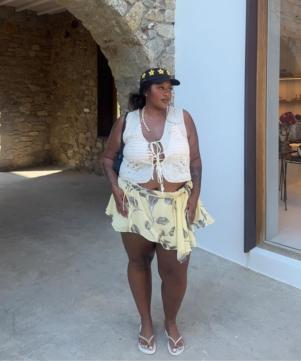 Modelo posa com colete branco de crochê aberto, saia amarela floral fluida e boné preto com estrelas, em clima leve.