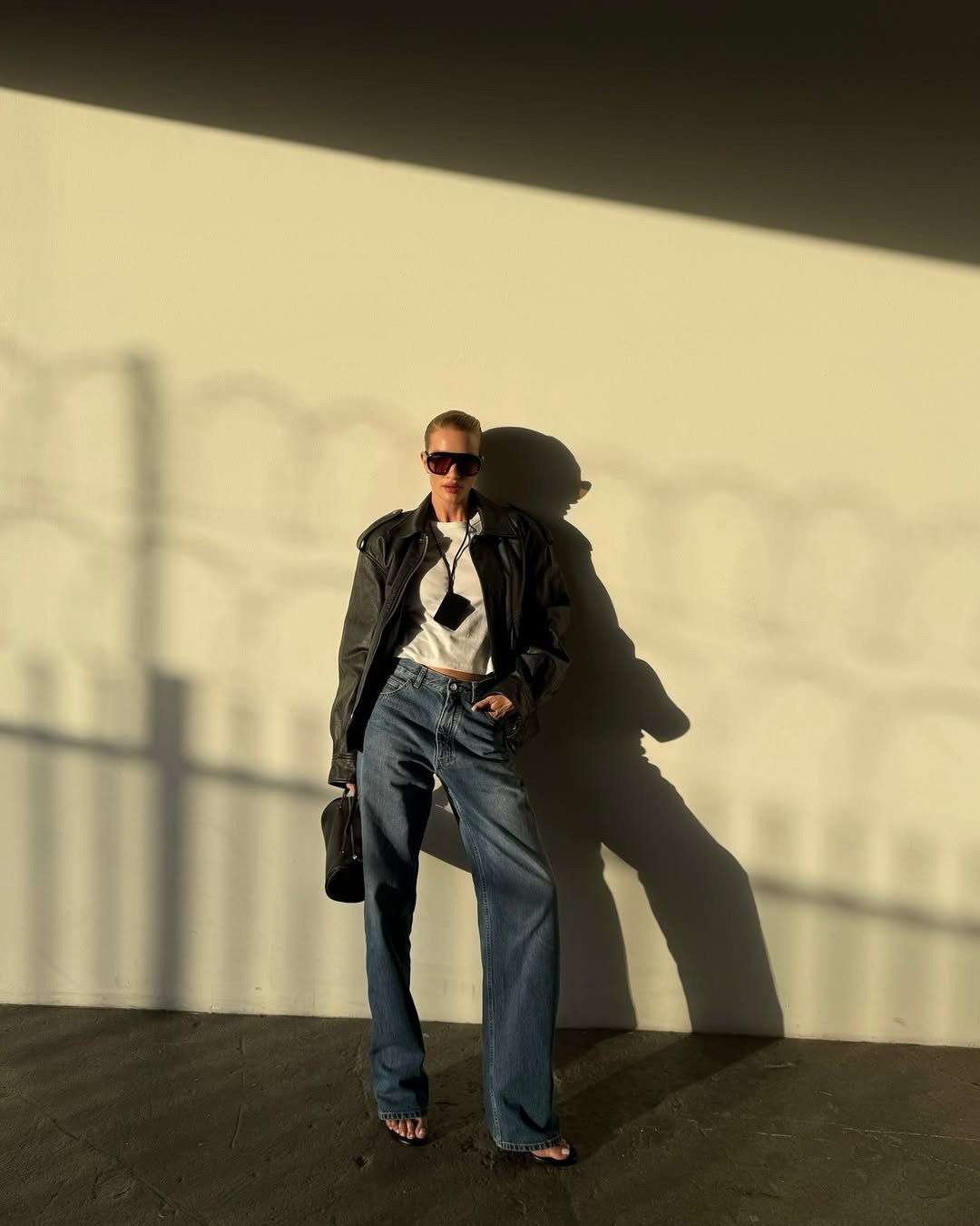 Modelo posa com colar funcional preto em cordão, jaqueta de couro oversized, camiseta branca e jeans azul, em clima urbano.