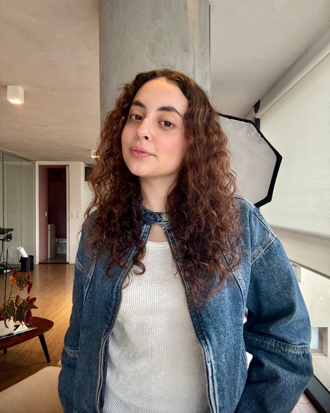 Mulher veste jaqueta jeans oversized e blusa branca de malha canelada, com resultado da rotina para cabelo cacheado por