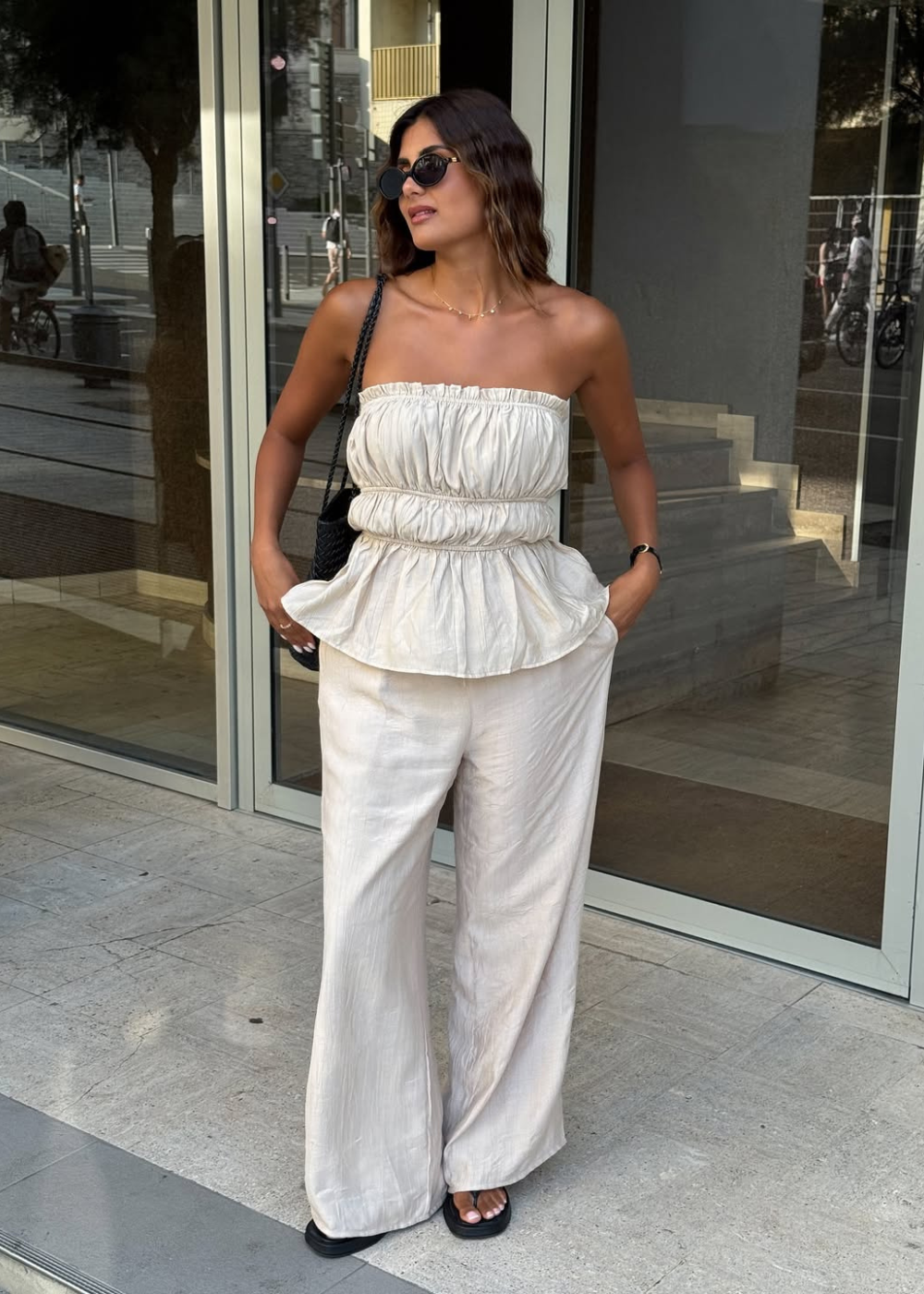 Mulher em pé usando roupas brancas bonitas, bolsa preta nos ombros e sandália aberta preta.