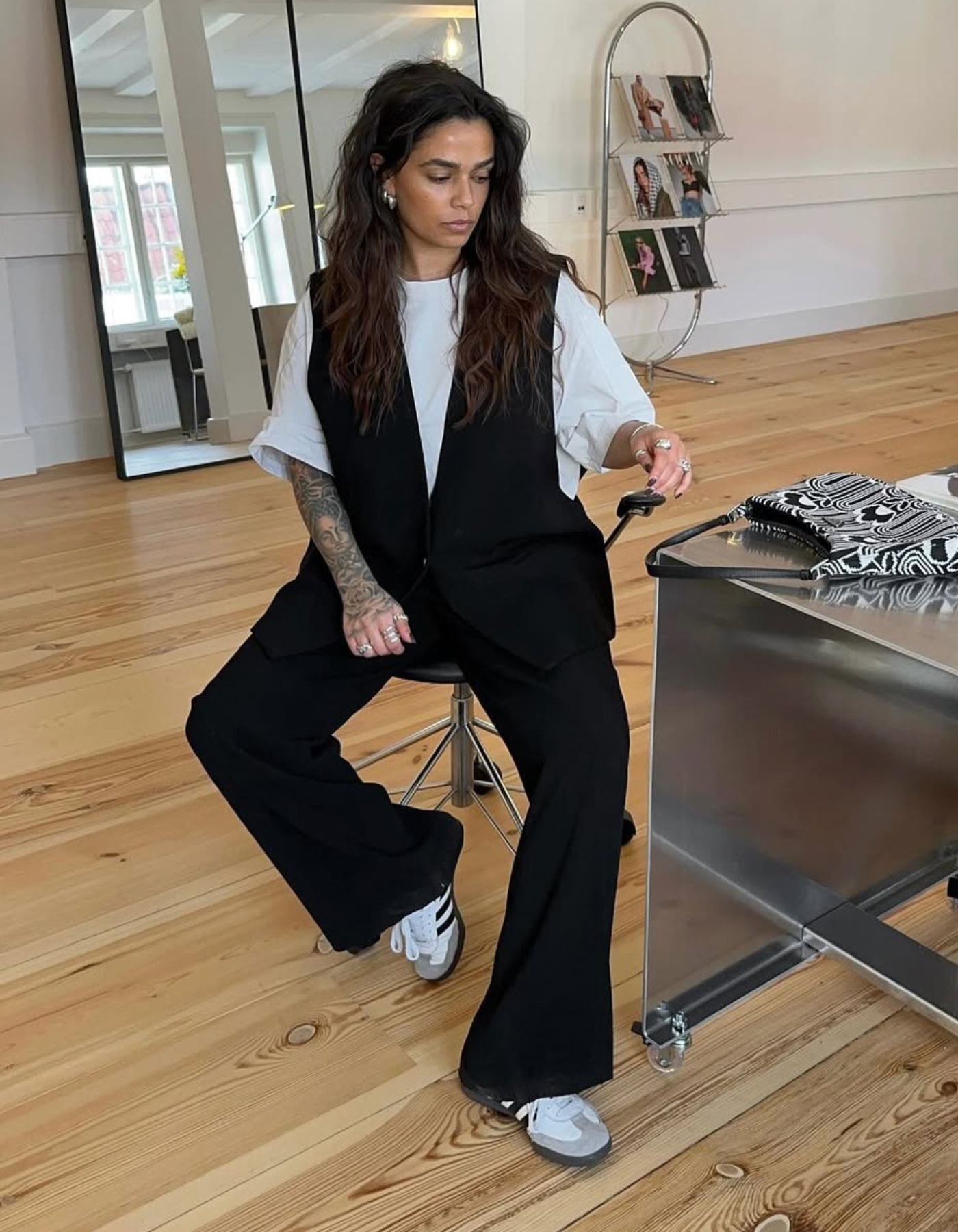 Modelo veste colete e calça preta de tecido fluido com camiseta branca larga e adidas sambae