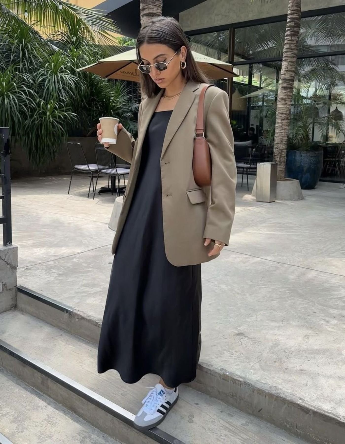 Modelo veste vestido preto longo fluido, blazer bege oversized e Adidas Sambae branco com listras