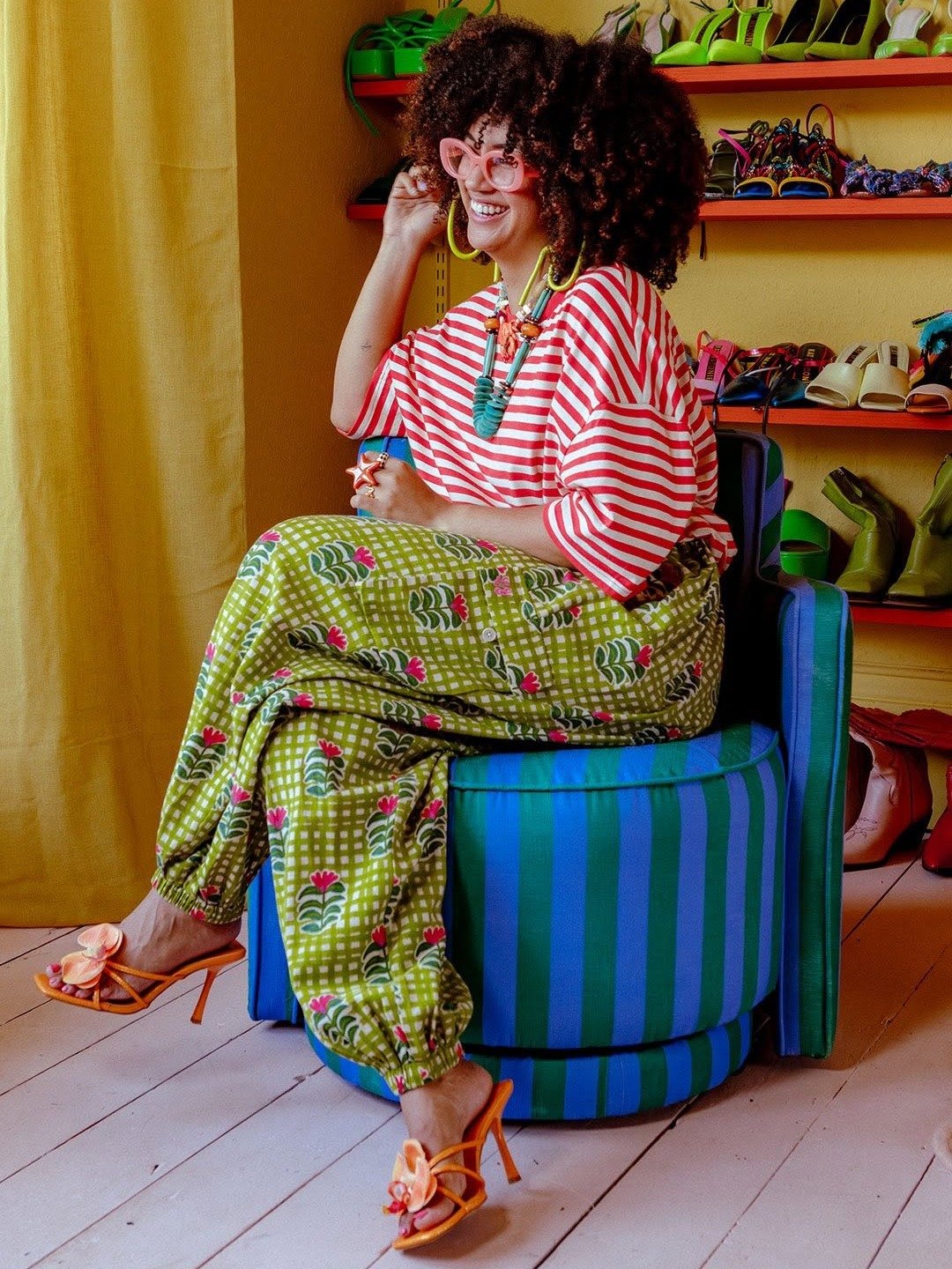 Mulher sentada sorri usando sandália de flor laranja com salto fino, calça estampada verde e blusa listrada vermelha.