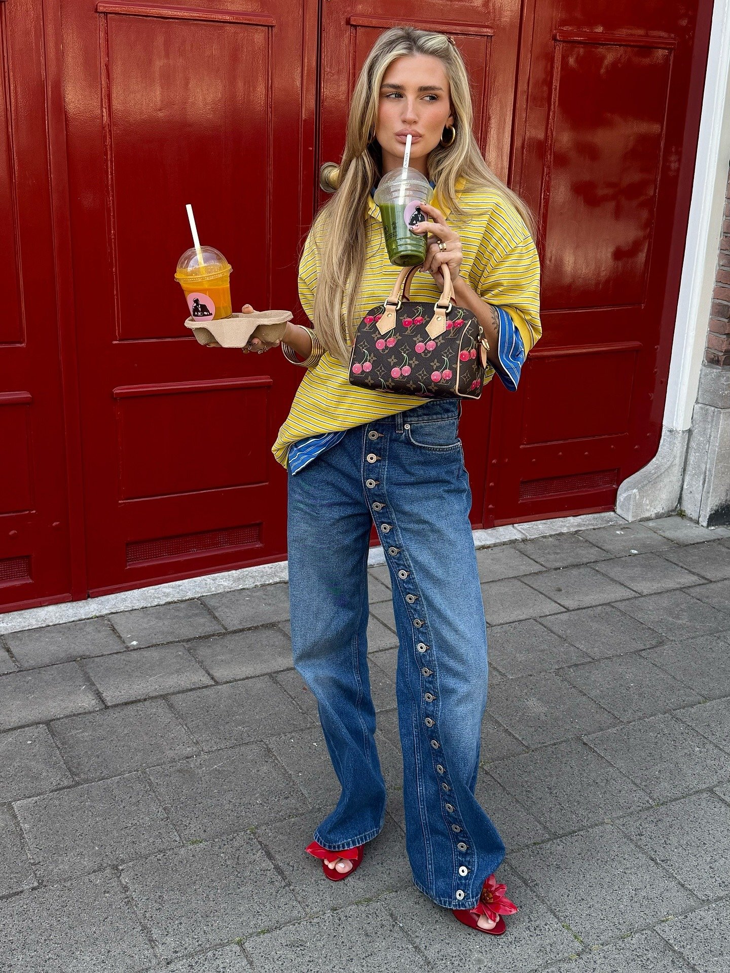 Modelo posa em frente a porta vermelha com blusa amarela listrada, jeans com botões laterais e sandália de flor vermelha.