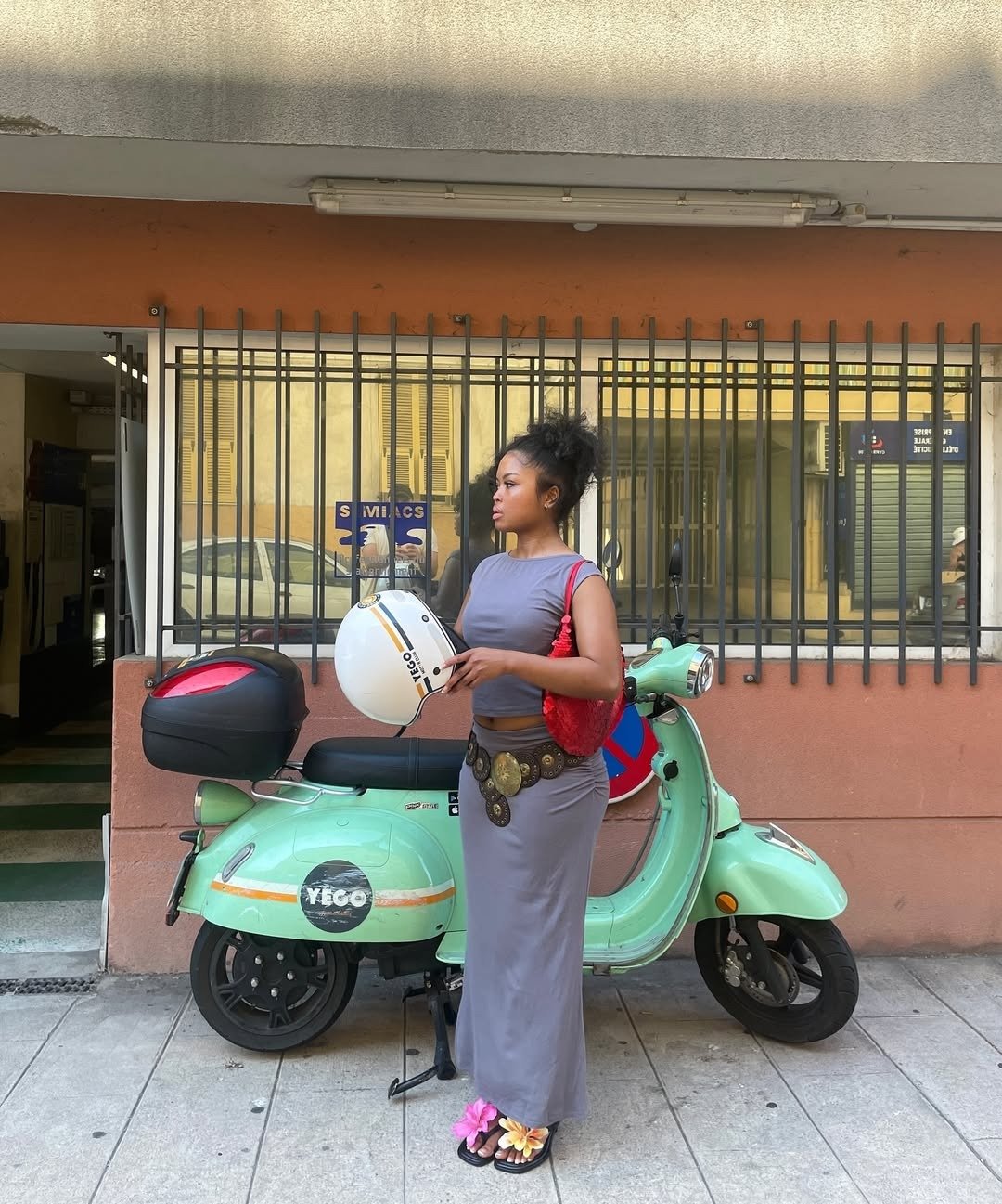 Mulher vestindo vestido cinza justo, cinto com medalhões, bolsa vermelha e sandália de flor.