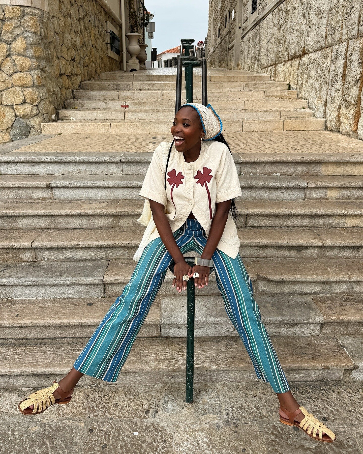 Modelo senta descontraída em escadaria usando sandálias da moda do verão 2026 em couro caramelo trançado, calça listrada