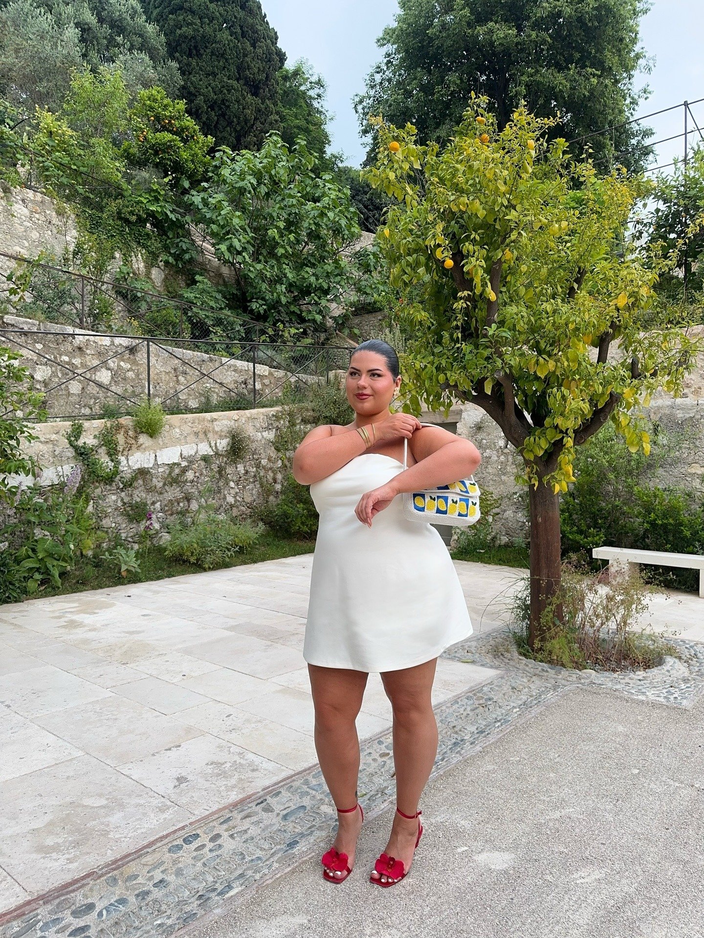 Modelo posa confiante com vestido branco curto e sandálias da moda do verão 2026 em vermelho vibrante com detalhes florais,