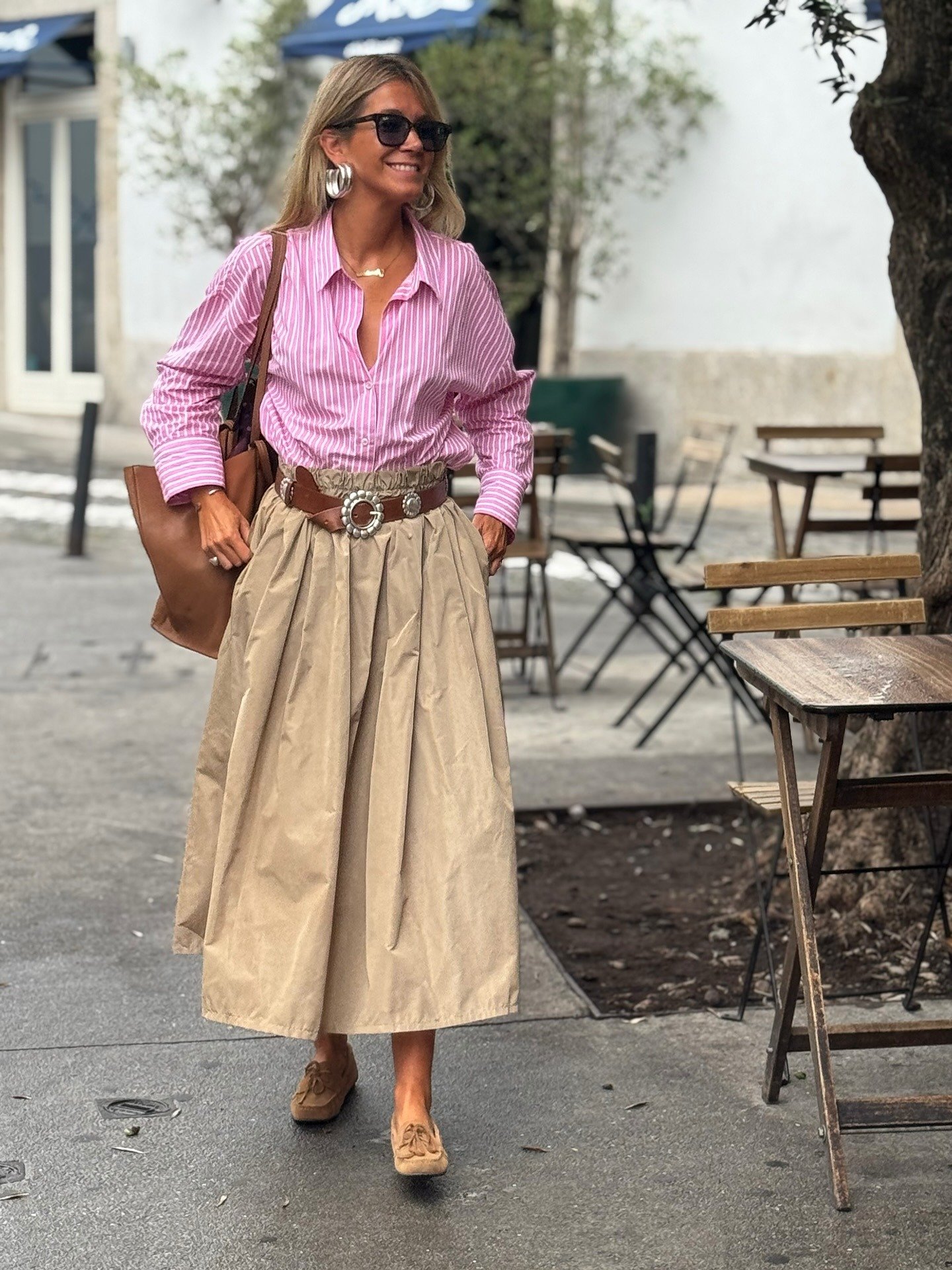 Mulher caminha sorrindo com sapato marrom de camurça, saia midi bege plissada, camisa rosa listrada e acessórios em prata.