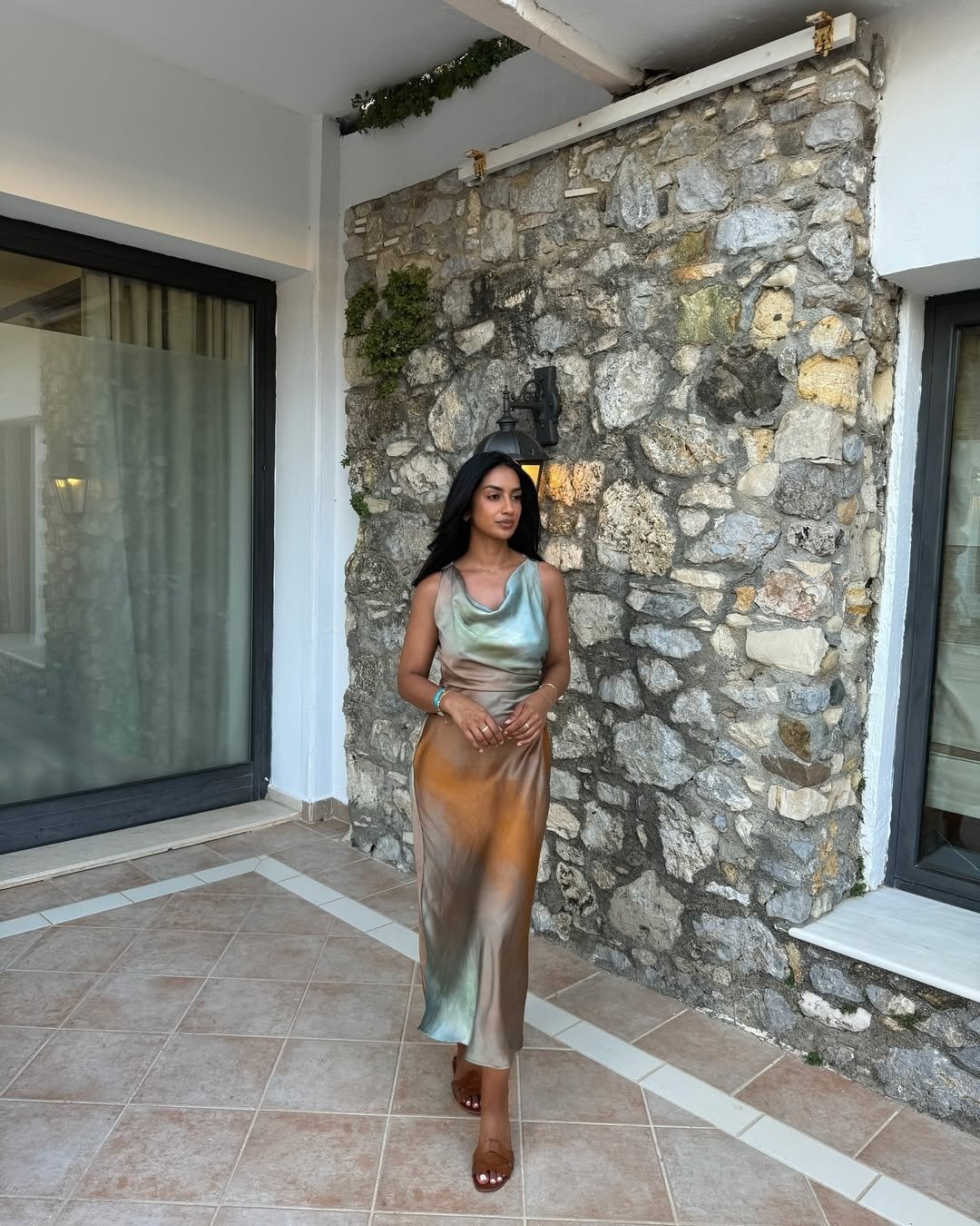 Modelo posa em vestido longo de tecido acetinado com tons terrosos e verdes, combinando com sapato marrom de tiras.