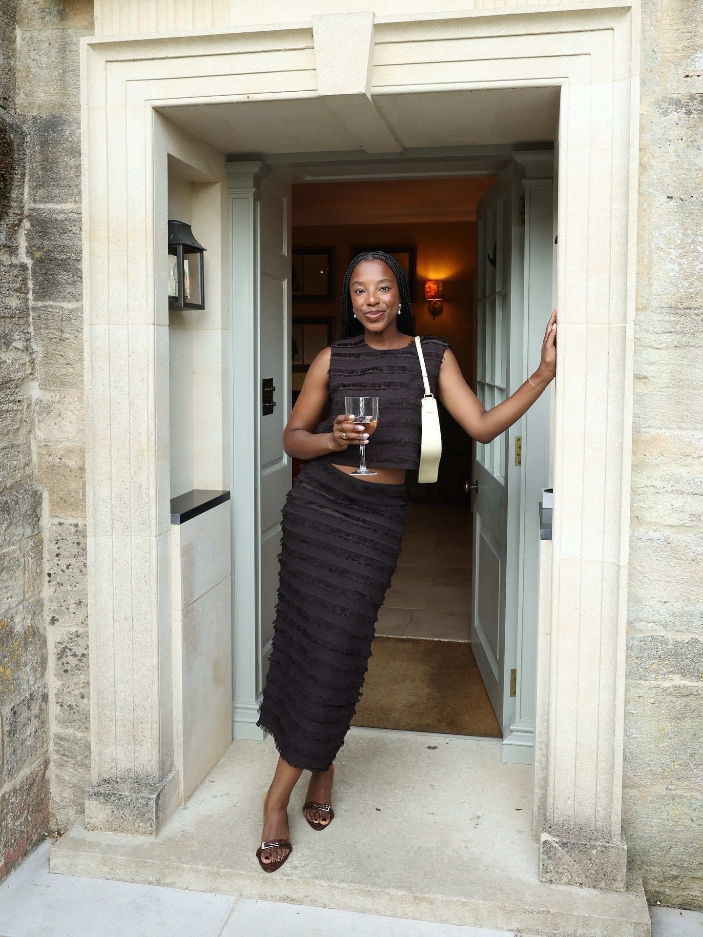 Modelo posa na entrada com vestido preto texturizado, bolsa branca e sapato marrom de salto médio com detalhe metálico.