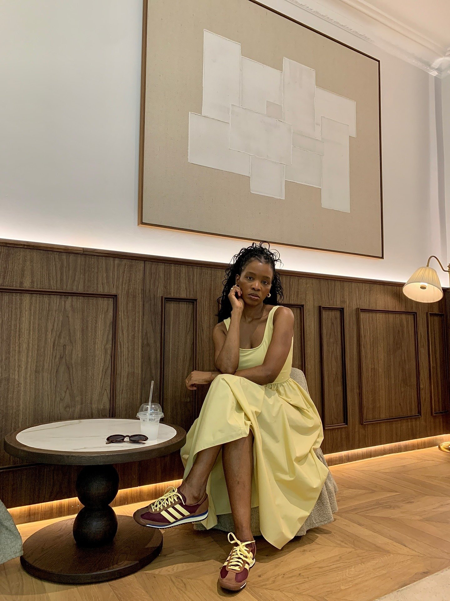 Modelo senta pensativa com vestido amarelo claro fluido e sapato marrom em couro e tecido, combinando estilo casual .