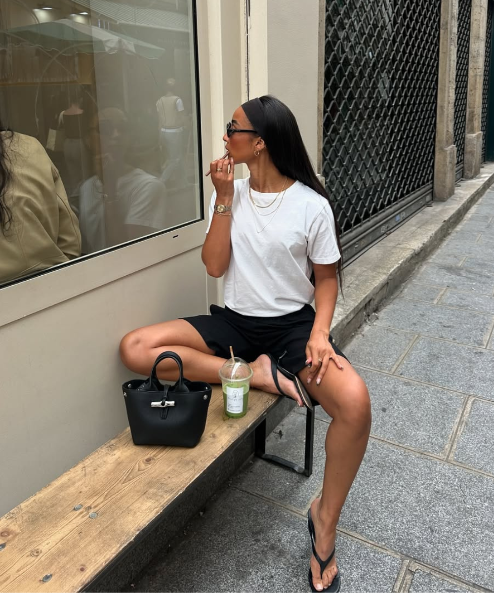 Modelo senta casualmente em banco de madeira na rua, usando sapatos femininos estilosos tipo chinelo preto com tiras finas,