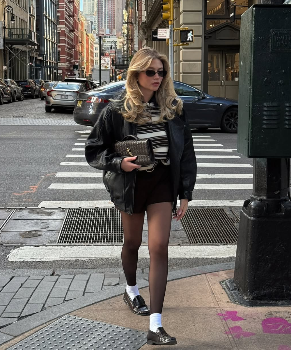 Modelo caminha na rua usando sapatos femininos estilosos tipo mocassim preto com meia branca, combinando jaqueta oversized