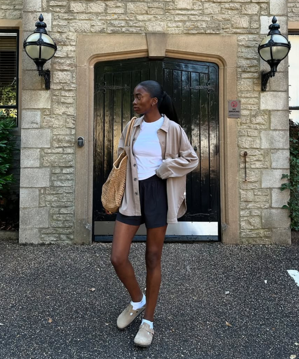 Modelo posa com sapatos femininos estilosos em camurça bege, combinados a meia branca, shorts pretos e camisa oversized