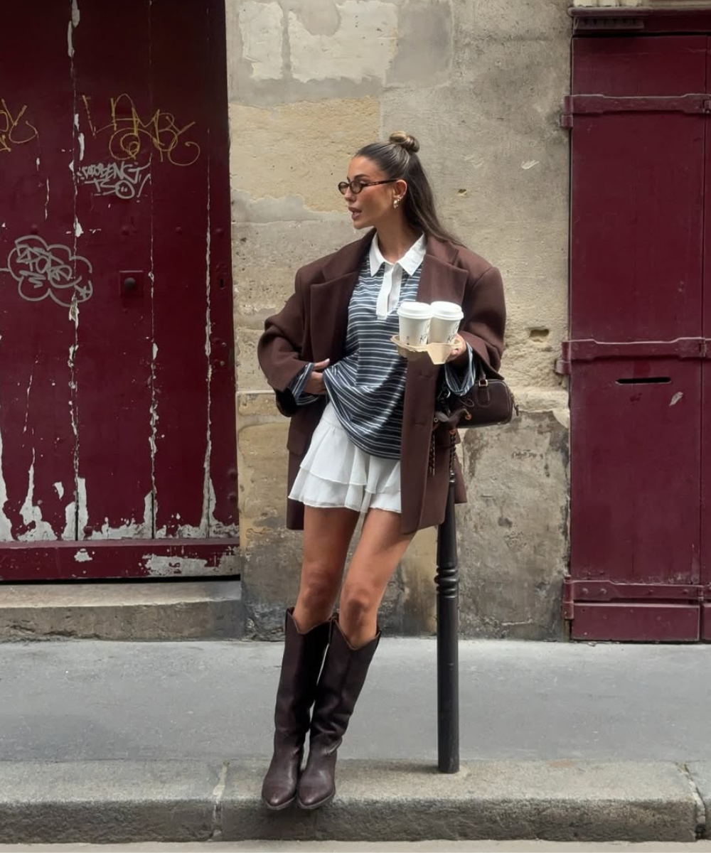 Modelo posa com blazer marrom oversized, camisa listrada, saia branca fluida e sapatos femininos estilosos em botas de couro