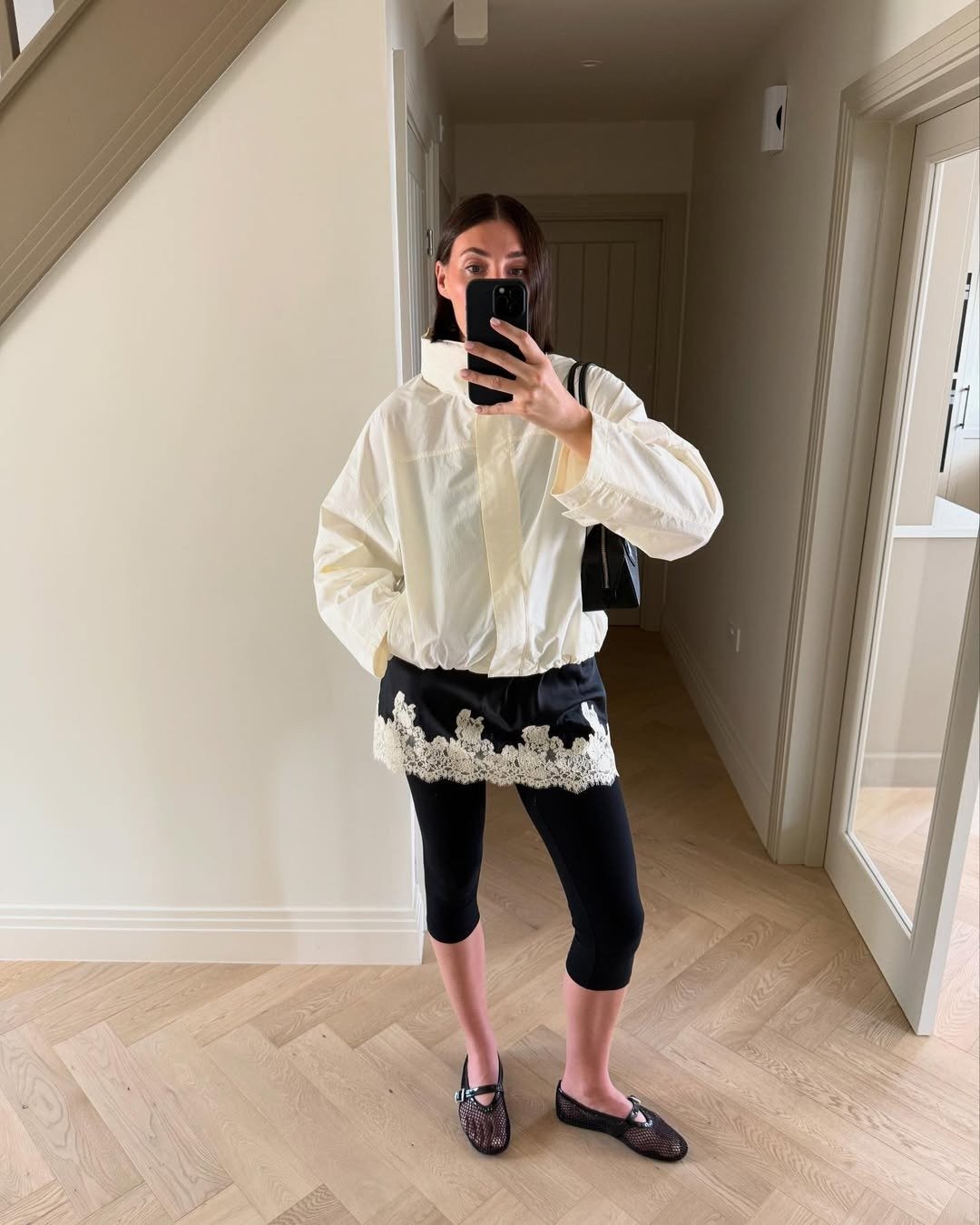 Modelo posa em ambiente interno com jaqueta off-white de gola funil estruturada, legging preta e saia curta com renda,