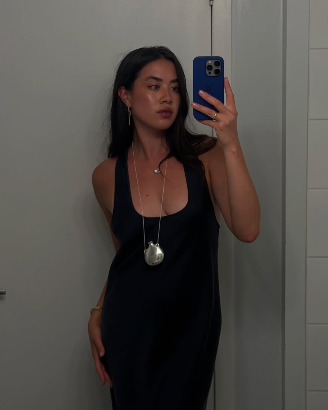 Modelo posa em vestido preto de alças largas com colar funcional longo em metal prateado, criando visual elegante e