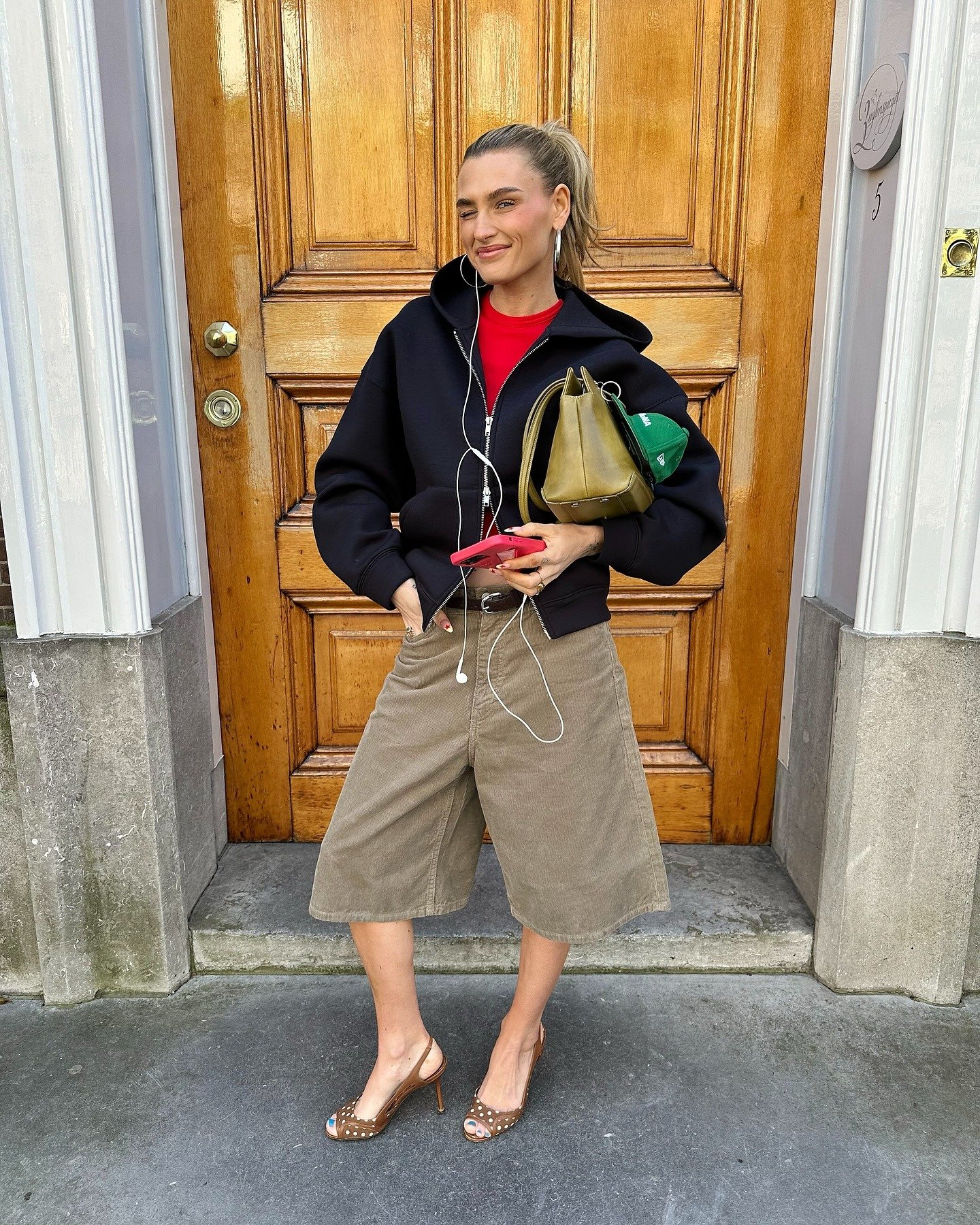 Modelo sorri confiante com scarpin clássico caramelo de salto fino e detalhes em poás, combinando bermuda ampla bege,