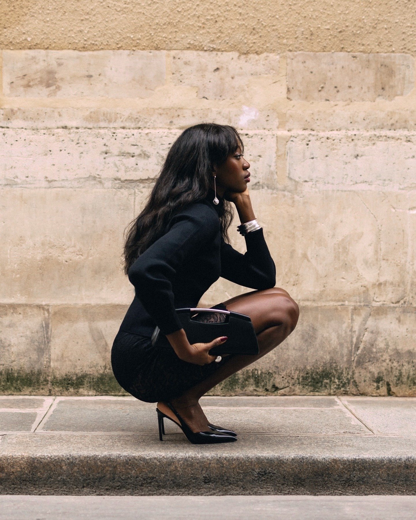 Mulher negra agachada em pose pensativa usa scarpin clássico preto de bico fino com vestido e suéter justos, transmitindo