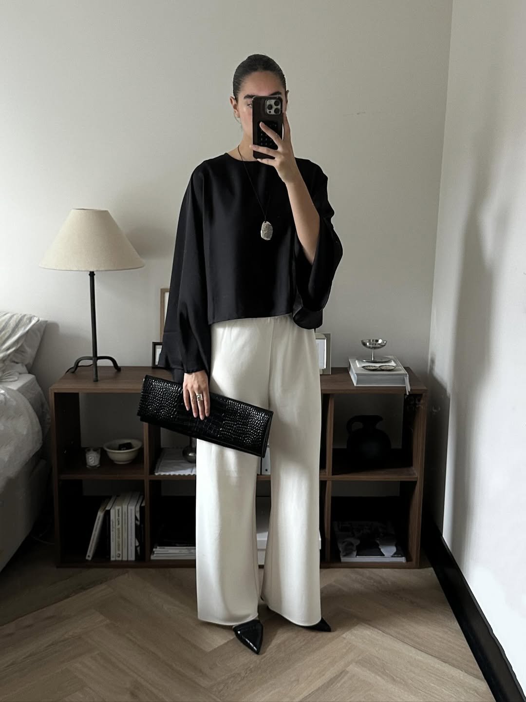 Modelo posa em ambiente minimalista com blusa preta de mangas amplas, calça branca ampla e colar funcional de pingente oval