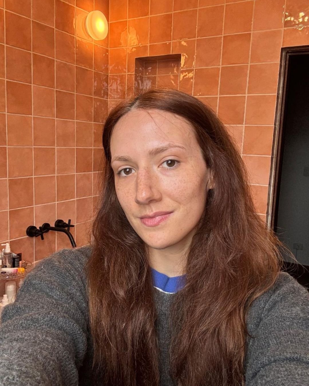 Em ambiente de banheiro com azulejos rosa, uma mulher com cabelo castanho e pele com sardas sorri depois da aplicação de sérum facial 