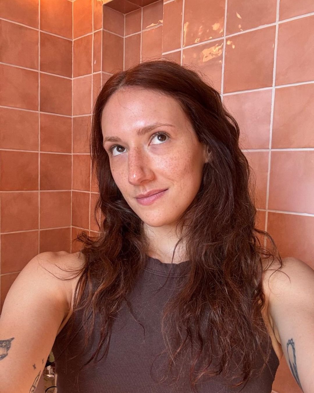 A imagem apresenta uma mulher com cabelo castanho ondulado, em um ambiente com azulejos em tons de rosa, sérum facial 