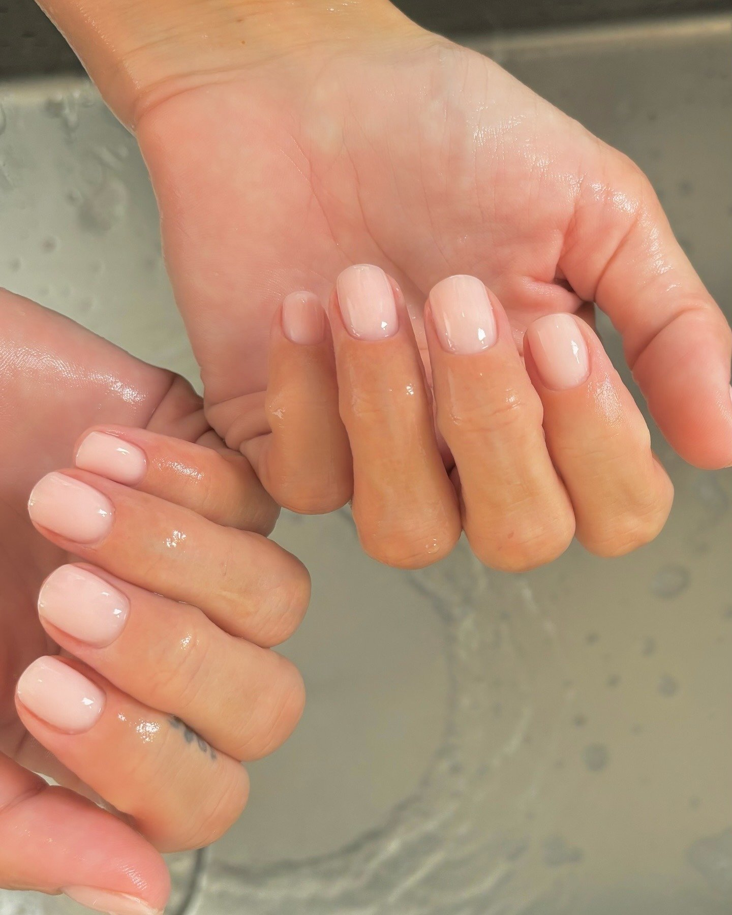 As unhas estão bem cuidadas, com um esmalte nude brilhante que destaca a suavidade e elegância das mãos. A imagem mostra duas mãos, uma em cima da outra, com gotas de água. Representa a tendência de soap nails.