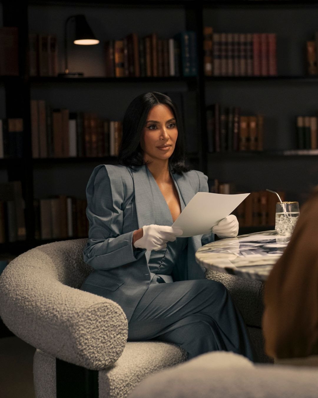 Cena de Tudo é Justo, série da Kim Kardashian, onde a personagem aparece em um ambiente sofisticado, sentada em uma poltrona arredondada, vestindo um elegante terno azul com ombreiras. Seu cabelo liso e maquiagem impecável destacam sua beleza.