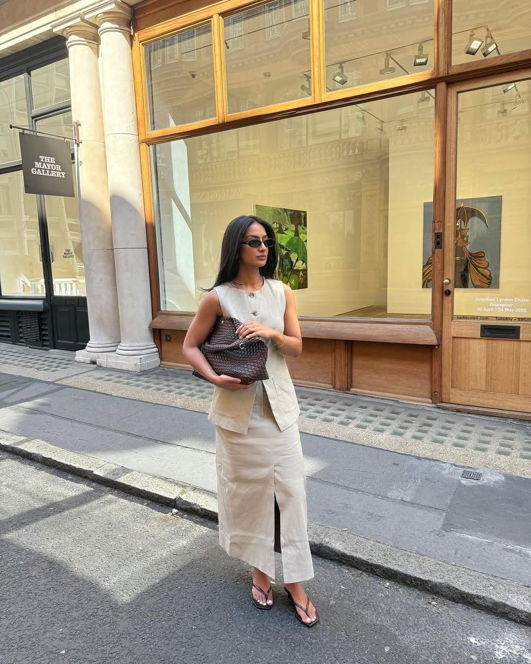 Modelo posa com tamanco de dedo preto minimalista, vestido bege de linho com fenda frontal e bolsa marrom trançada, em