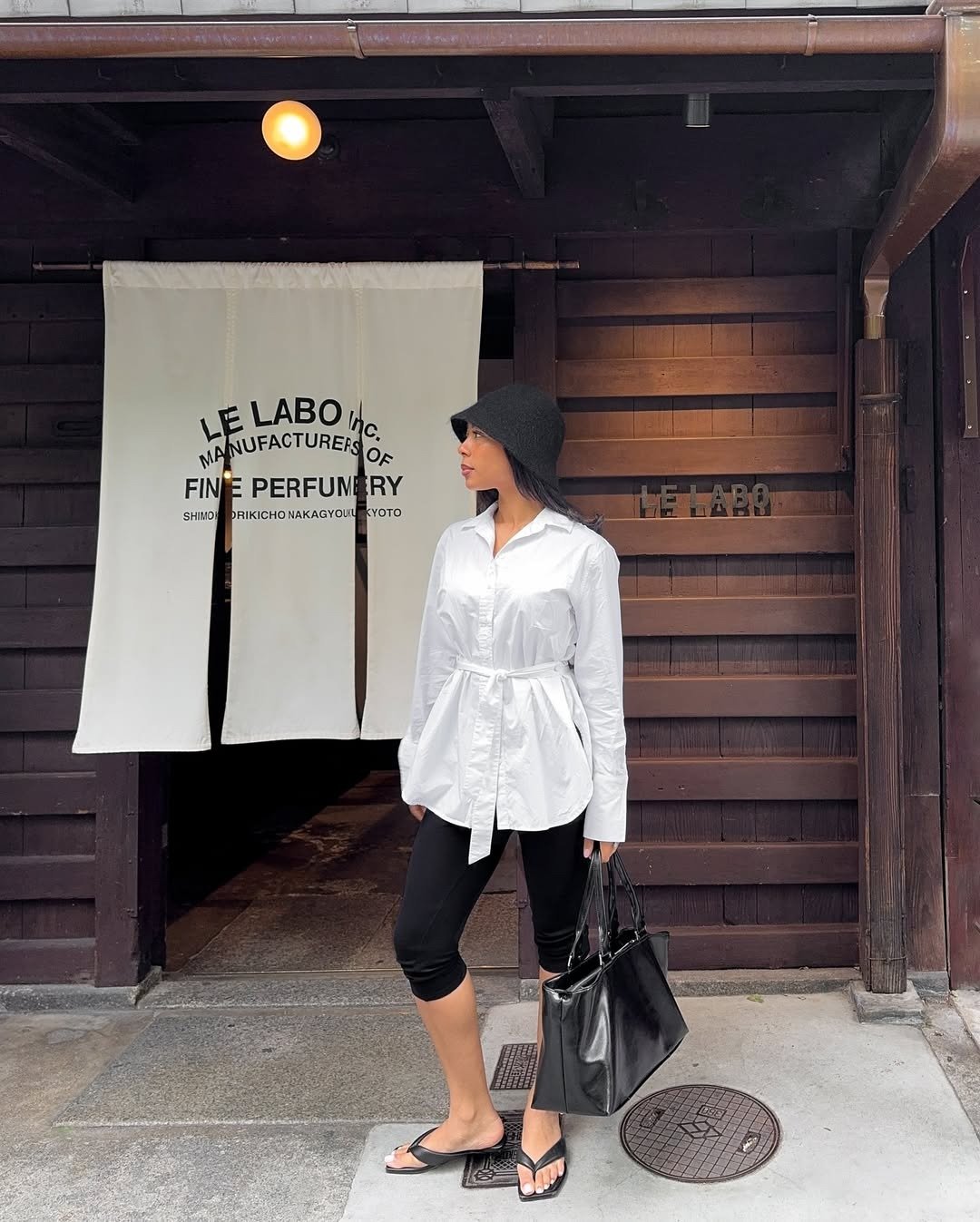 Modelo posa com tamanco de dedo preto em material sintético, combinando com legging preta e camisa branca oversized, em