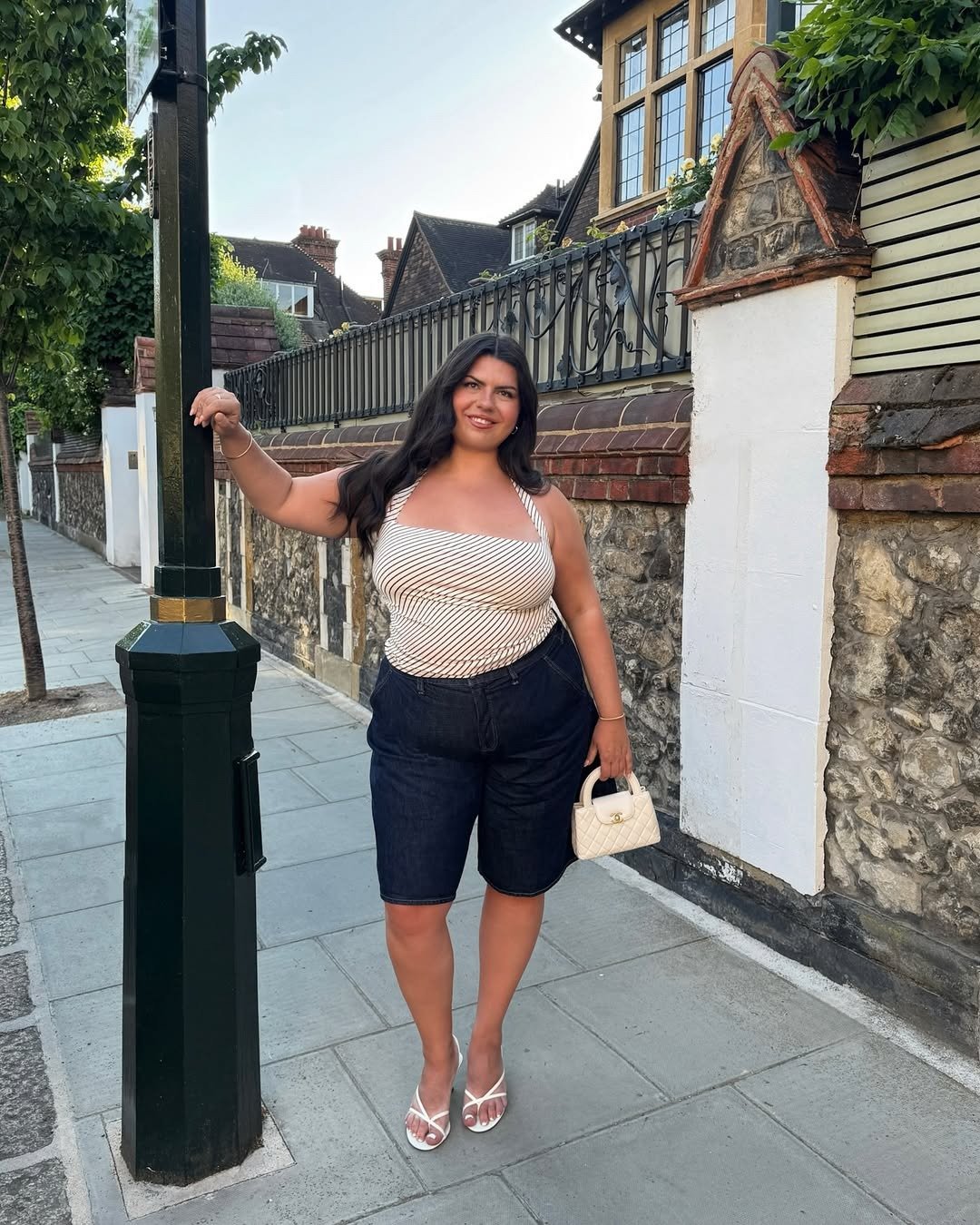 Modelo posa sorridente com tamanco de dedo branco em tiras finas, bermuda jeans escura e blusa listrada, em cenário urbano